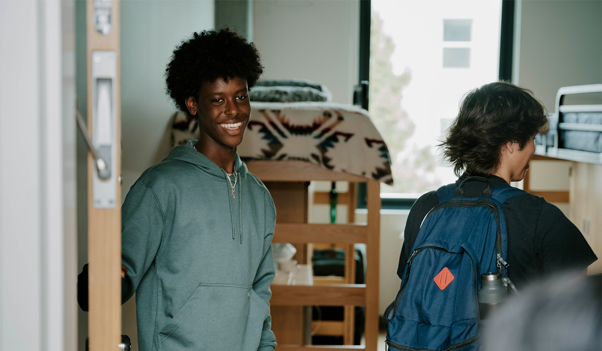 A student smiles while holding the door open to his room in the West Grove Commons while another person wearing a backpack walks into the room two with loft beds and a window visible behind them